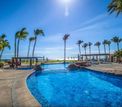 Crystal-blue oceanfront resort pool with rock features and cabanas, palm trees swaying under a sunny sky overlooking the beach.