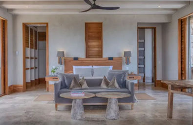 Sunny modern coastal bedroom suite with king bed, gray loveseat and textured pillows, two round stone pedestal coffee tables, wooden headboard with bedside lamps, ceiling fan and wide wood‑framed openings to a patio.