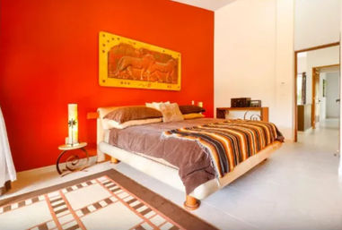 Bright bedroom with bold red accent wall, low platform bed dressed in brown bedding and a striped orange-and-brown throw, carved elephant wall art above, patterned rug, bedside lamp, and open doorway to a light hallway.