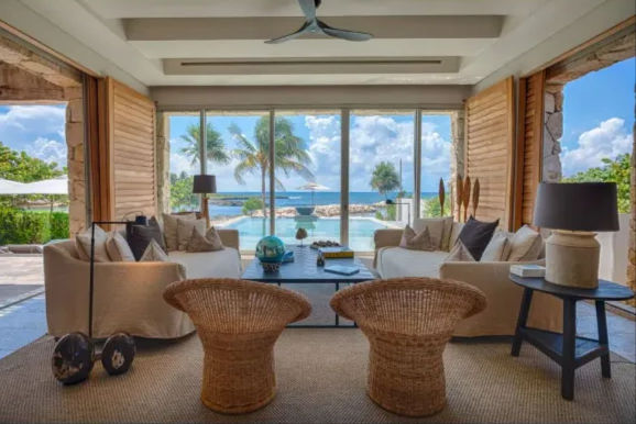 Sunlit beachfront living room in a tropical villa with wicker chairs and neutral sofas around a coffee table, floor-to-ceiling glass opening to a pool, palm trees and turquoise ocean under a blue sky