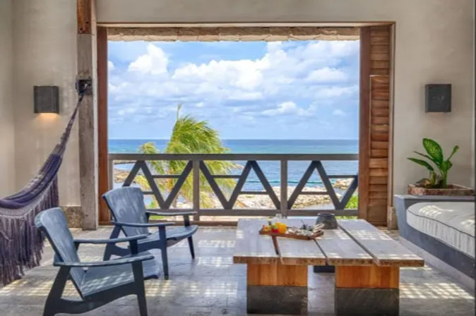 Cozy tropical beachfront bungalow interior with hammock, blue chairs and low wooden table opening onto a balcony overlooking turquoise ocean and a swaying palm.