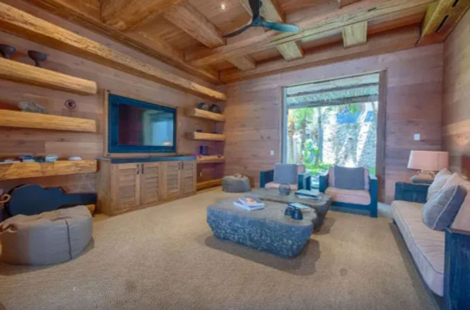 Cozy rustic wood-paneled tropical cottage living room with built-in shelves and TV, stone coffee table, cushioned sofa and chairs, ceiling fan, and sliding door to a palm-lined garden.