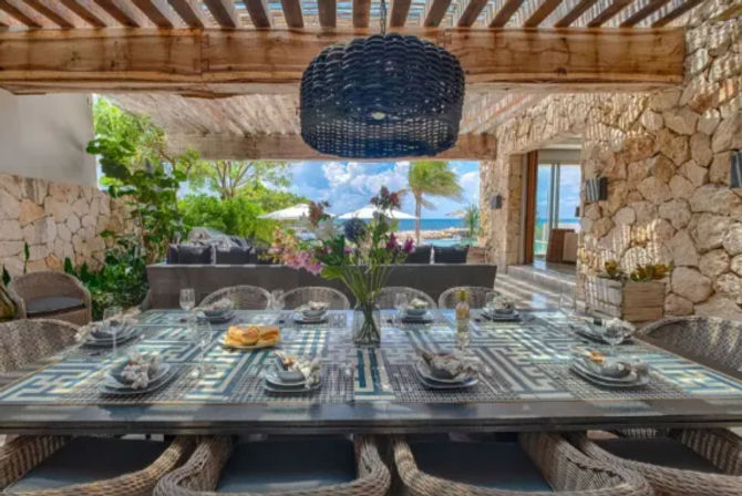 Oceanfront covered patio at a tropical villa — large patterned dining table set for eight with wicker chairs, woven pendant light, stone walls, lush plants, and palm trees, umbrellas and turquoise sea visible beyond.