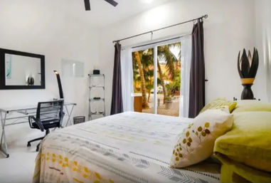 Bright modern bedroom with green-and-yellow bedding, sliding glass doors opening to palm trees and a sandy tropical courtyard, desk and office chair, and ceiling fan