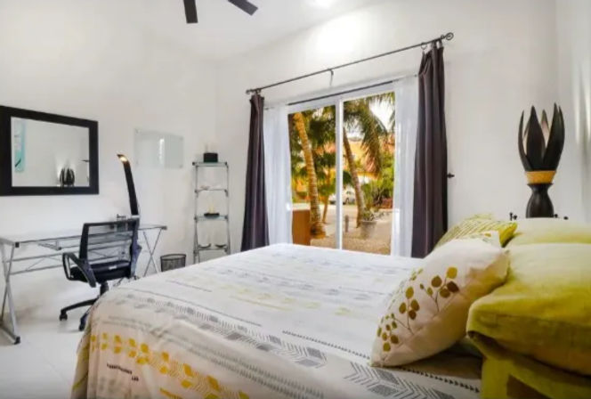 Bright modern bedroom with green-and-yellow bedding, sliding glass doors opening to palm trees and a sandy tropical courtyard, desk and office chair, and ceiling fan