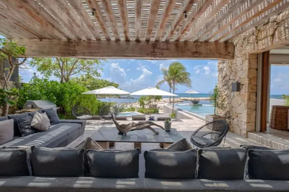 Tropical oceanfront patio under a slatted wooden pergola with gray lounge sofas, coffee table, wicker chairs, an infinity pool, white umbrellas and a palm tree overlooking turquoise sea.