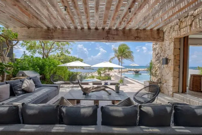 Tropical oceanfront patio under a slatted wooden pergola with gray lounge sofas, coffee table, wicker chairs, an infinity pool, white umbrellas and a palm tree overlooking turquoise sea.