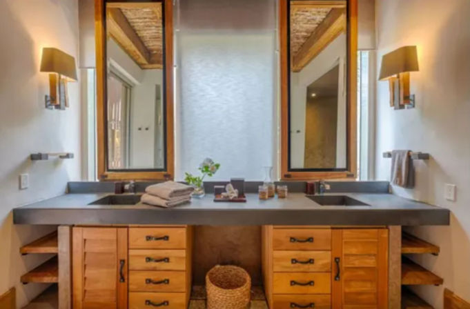 Spa-like double vanity bathroom with matching wood cabinets and drawers, dark concrete countertop with two inset sinks, tall framed mirrors flanking a frosted window, wall sconces, folded towels and a woven basket underneath.