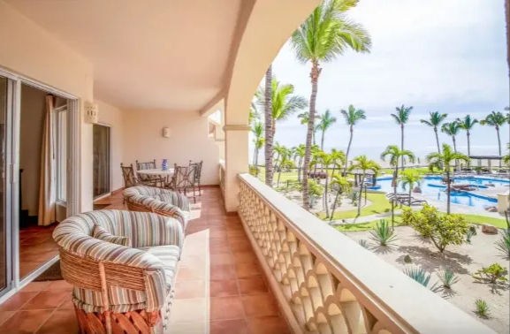 Oceanfront resort balcony with terracotta tiles, striped wicker lounge chairs and dining set, arched railing overlooking palm trees, landscaped grounds, pools and beach