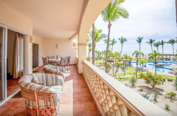 Oceanfront resort balcony with terracotta tiles, striped wicker lounge chairs and dining set, arched railing overlooking palm trees, landscaped grounds, pools and beach