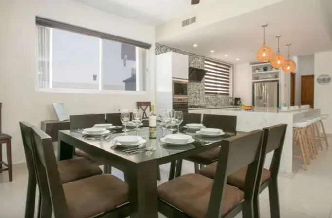 Bright modern open-concept dining and kitchen with dark wood table set for six, wine and glassware, white island, bar stools, pendant lights and mosaic backsplash.
