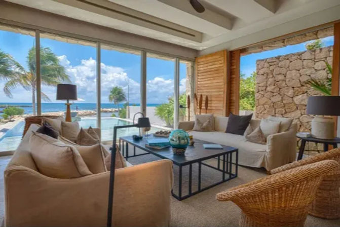 Sunlit beachfront living room with neutral sofas, rattan chairs and a coffee table, floor-to-ceiling windows overlooking a pool, palm trees and ocean view.