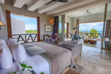 Sunlit oceanfront bedroom with king bed in gray linens, sliding doors to a covered terrace with hammock, lounge seating and wooden railing overlooking turquoise sea and palm trees.