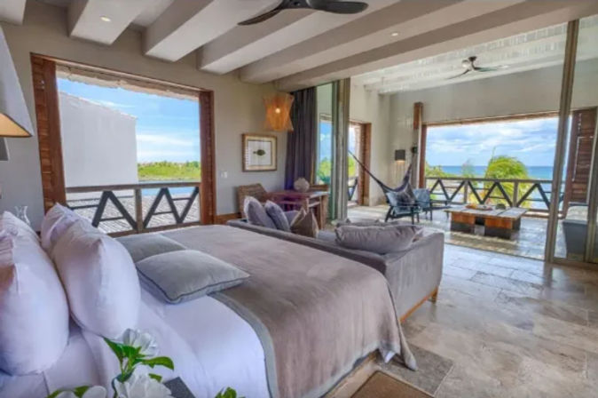 Sunlit oceanfront bedroom with king bed in gray linens, sliding doors to a covered terrace with hammock, lounge seating and wooden railing overlooking turquoise sea and palm trees.
