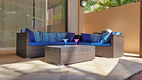 Sunlit outdoor patio with brown rattan L-shaped sectional, bright blue cushions and striped pillows, wicker coffee table topped with blue and pink cocktail glasses, beige wall and palm shadows.
