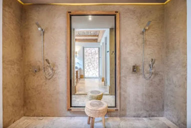 Spa-like walk-in double shower with textured beige plaster walls, two wall-mounted showerheads, large framed mirror reflecting a stone feature wall and hallway, and a woven round stool on a tiled floor.