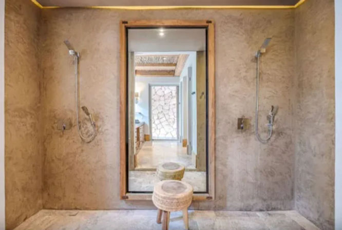 Spa-like walk-in double shower with textured beige plaster walls, two wall-mounted showerheads, large framed mirror reflecting a stone feature wall and hallway, and a woven round stool on a tiled floor.