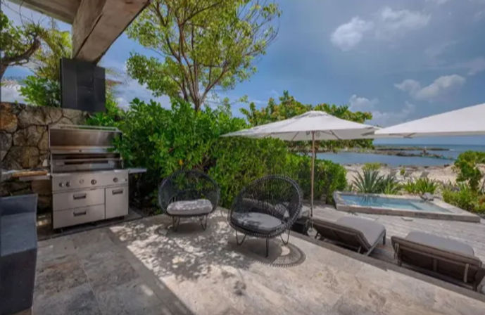 Oceanfront stone patio with stainless steel outdoor grill and round wicker chairs under a sun umbrella; chaise lounges and a small plunge pool overlook a sandy beach and turquoise sea.