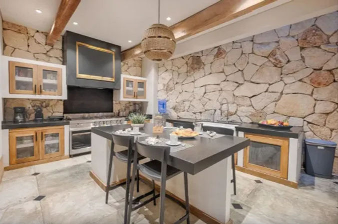Cozy rustic kitchen with textured stone wall, central island and black countertop with bar stools, woven pendant light, exposed wooden beams, stainless range and glass-front cabinets set for breakfast.