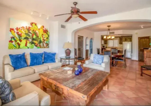 Bright Southwestern-style open-plan living room with beige sofas and blue pillows, rustic wooden coffee table on terracotta tile floor, colorful cactus wall art, ceiling fan, and arched view into the dining area and kitchen