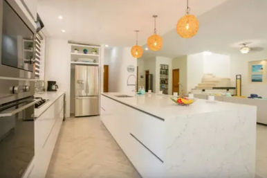 Bright modern open-plan kitchen with a large white marble island, stainless steel appliances, and three woven pendant lights above, opening to a living area.