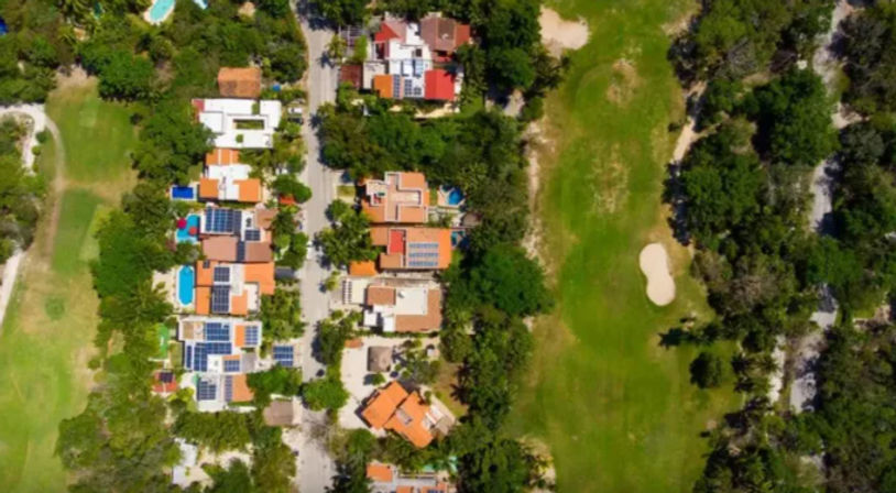 Aerial drone view of a residential neighborhood with orange-tile roofs, backyard pools and solar panels along a tree-lined street beside a green golf fairway and sand bunker.