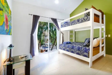 Bright tropical vacation-style bedroom with white bunk beds and blue floral bedding, green accent wall, sliding glass door to a palm-lined balcony, gray curtains and glass side table