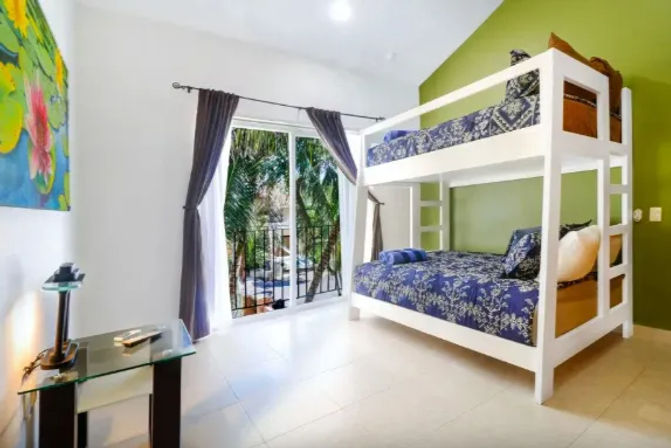 Bright tropical vacation-style bedroom with white bunk beds and blue floral bedding, green accent wall, sliding glass door to a palm-lined balcony, gray curtains and glass side table