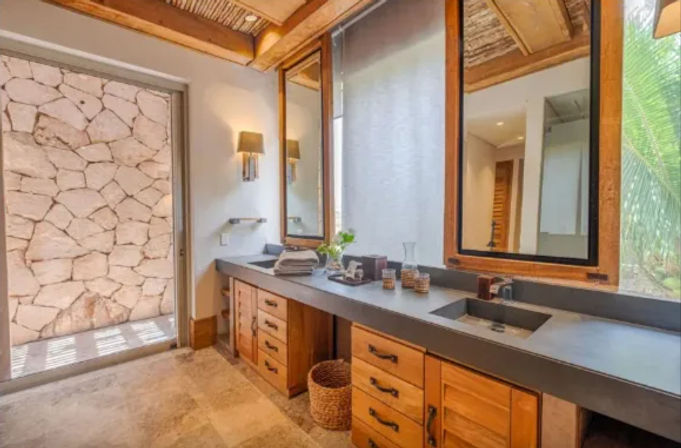 Spa-like tropical bathroom with long concrete double vanity, wooden cabinetry, large mirrors, stone accent wall and palm-framed window