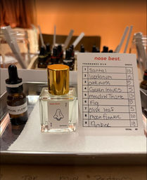 Square glass perfume bottle with gold cap and nose illustration on a metal tray, next to a dropper bottle labeled “moon flower” and a handwritten fragrance oils card listing santal, labdanum, oakmoss, green leaves, meadow thistle, fig, olive leaf, moonflower, amber — perfume blending setup with scent strips and amber vials.