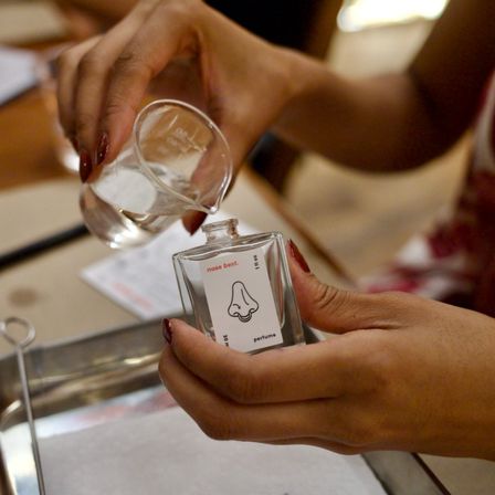 Close-up of hands pouring liquid from a small glass beaker into a perfume bottle labeled "nose best" during a fragrance blending workshop