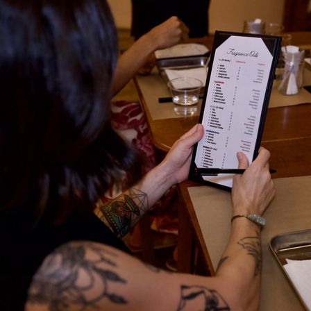 Person with tattooed arms holding a framed "Fragrance Oils" menu at a wooden table, pen in hand with glass, trays and notepad nearby