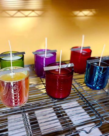Colorful glass jar candles with centered wicks cooling on a metal wire rack against a warm yellow background