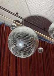 Large mirrored disco ball with smaller mirror balls hanging from chains in an indoor event space, casting dotted light shadows on a patterned tin ceiling above red velvet curtains.