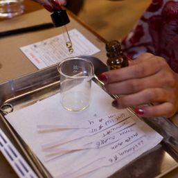 Close-up of hands using a glass dropper to add essential oil into a small beaker on a metal tray, with labeled scent testing strips and handwritten notes at a scent-testing station.