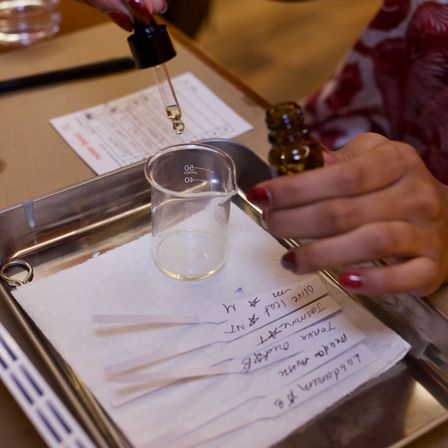 Close-up of hands using a glass dropper to add essential oil into a small beaker on a metal tray, with labeled scent testing strips and handwritten notes at a scent-testing station.