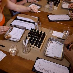 Fragrance workshop table with hands sampling scent strips, wooden rack of essential oil bottles, glass jars, metal trays of labeled strips, sample cups and printed scent menus.