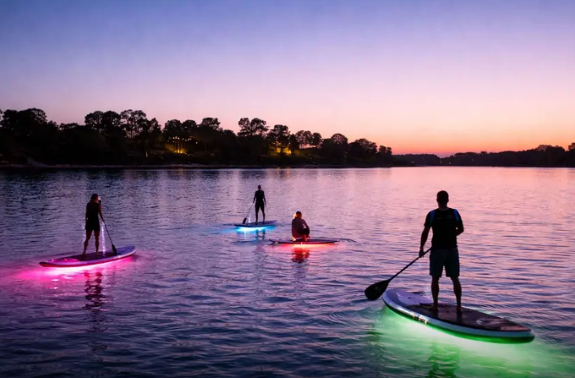 Four stand-up paddleboarders silhouetted against a purple-orange sunset on a calm lake, each on LED-lit boards glowing pink, blue, red and green near a tree-lined shore.