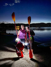 Two smiling adults ready for night kayaking, holding red paddles and lanterns on a sandy lakeshore at dusk with calm water and silhouetted mountains in the background.