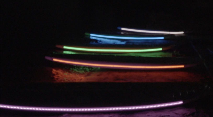Colorful LED-lit surfboards lined up on a dark beach at night, glowing purple, orange, green and blue with reflections on wet sand.