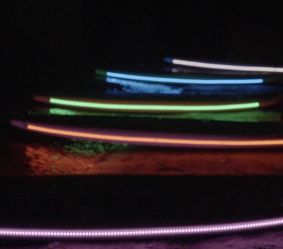 Colorful LED-lit surfboards lined up on a dark beach at night, glowing purple, orange, green and blue with reflections on wet sand.
