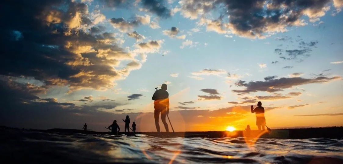 Silhouettes of paddleboarders and beachgoers at a golden coastal sunset, dramatic cloud-filled sky and gentle ocean waves reflecting warm light.