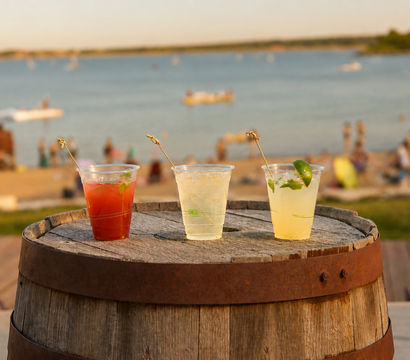 Three colorful summer cocktails in plastic cups with garnishes on a wooden barrel, seaside beach and boats on a calm bay at sunset