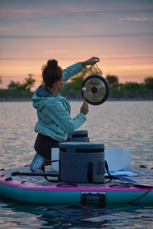 Person kneeling on a pink-and-teal paddleboard at sunset on a calm lake, holding a gong and mallet for a peaceful sound‑healing session