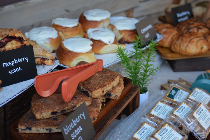 Bakery market display with frosted cinnamon rolls, raspberry and blueberry scones, flaky croissants, orange tongs and packaged snack bars on wooden trays with a small potted herb garnish.