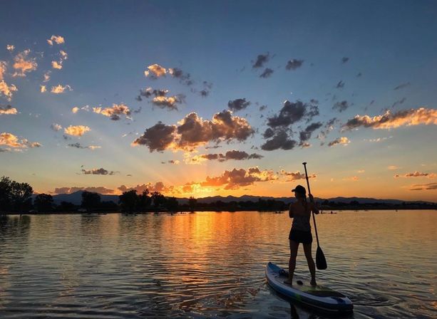 Silhouette of a person paddling a stand-up paddleboard on a calm mountain lake at sunset, golden sky, scattered clouds and warm reflections on the water