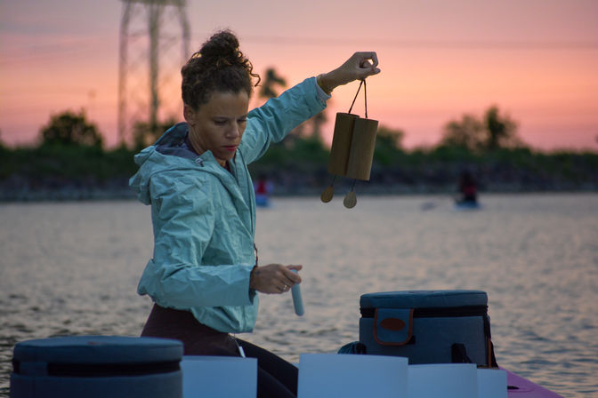 Person seated on a kayak at sunset holding wooden chimes over calm water, soft pink sky and tree-lined shore in the background.