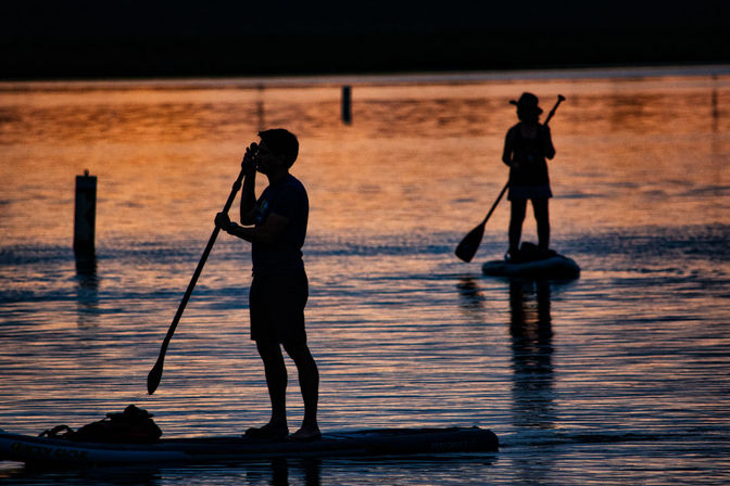 Two stand-up paddleboarders silhouetted on a calm lake at sunset, golden-orange reflections rippling across the water.