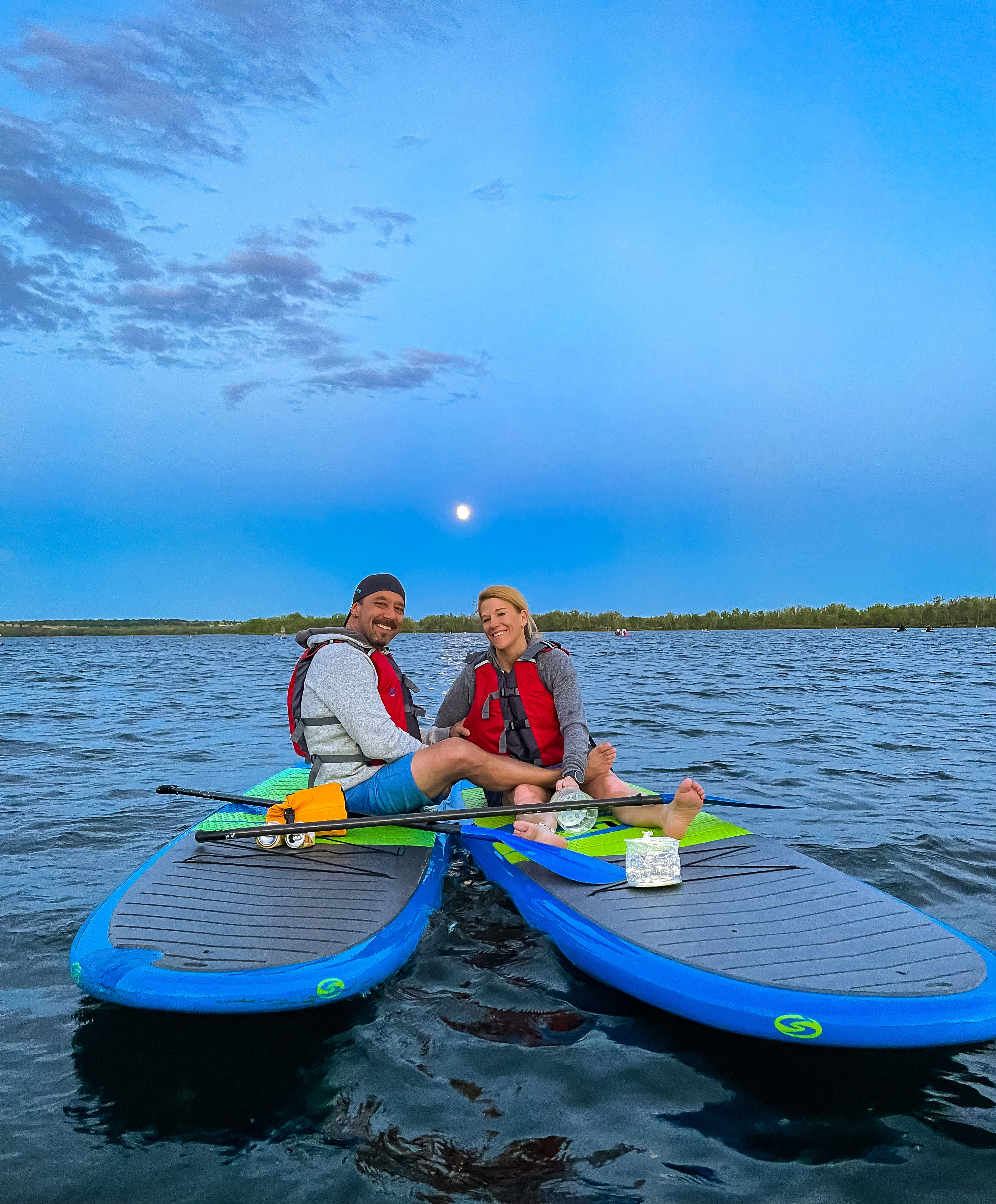 Two people in red life jackets sitting cross-legged on blue paddleboards, smiling on a calm lake at dusk with a full moon rising above a tree-lined horizon