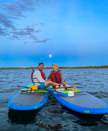 Two people in red life jackets sitting cross-legged on blue paddleboards, smiling on a calm lake at dusk with a full moon rising above a tree-lined horizon